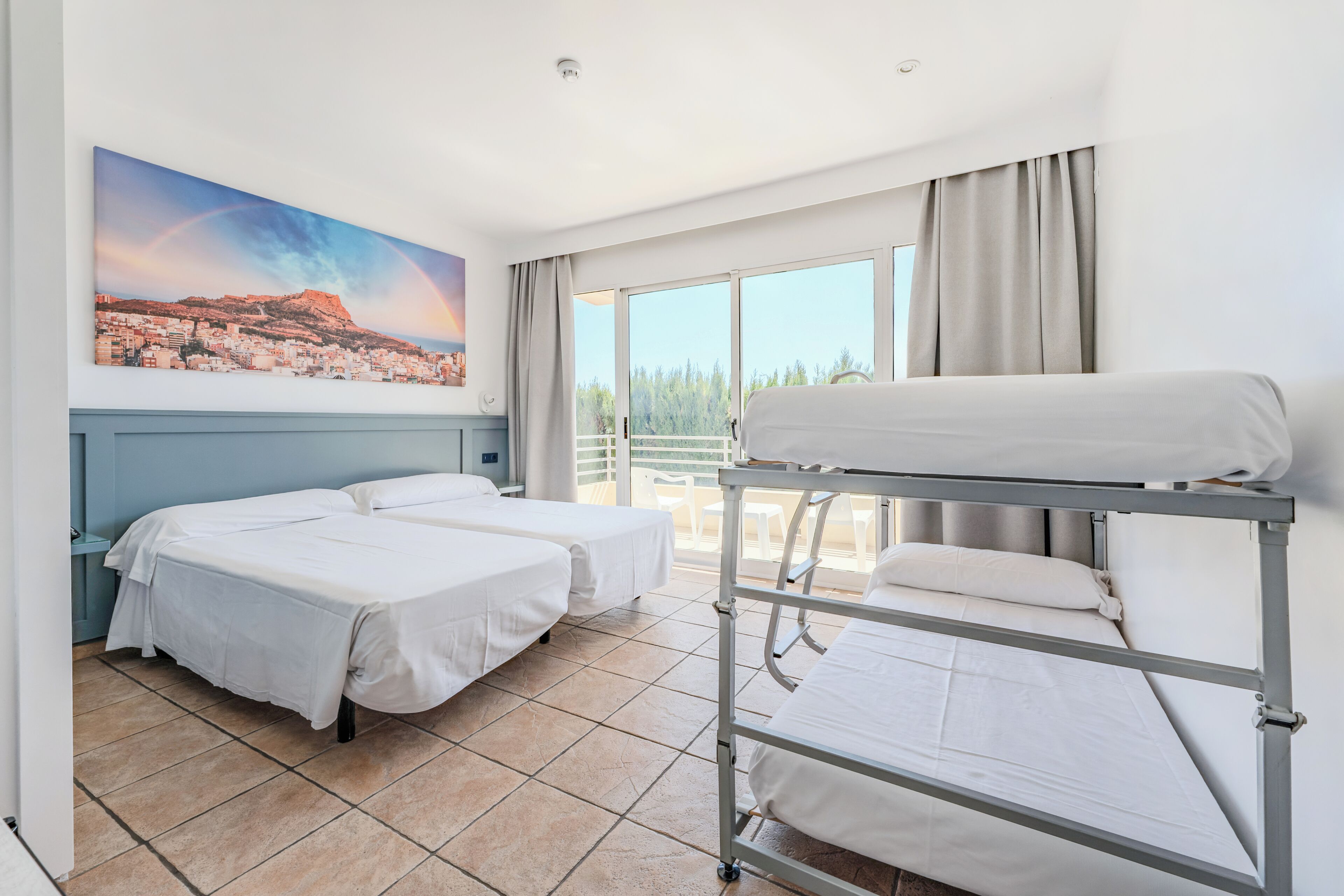 A hotel room with two single beds, a bunk bed, a large window with a view of a cityscape, and a painting of a rainbow over a city.