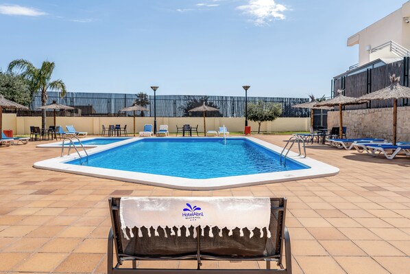 Une piscine entourée de chaises longues et de parasols, avec une terrasse carrelée et un bâtiment en arrière-plan.