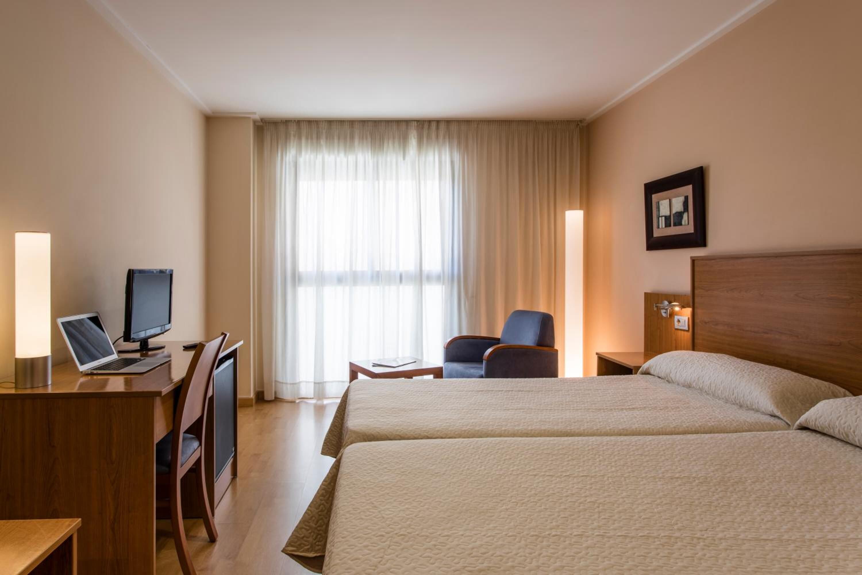 A hotel room with two beds, a desk with a laptop and a monitor, a chair, and a window with curtains.