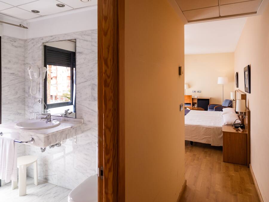 A hotel room with a bathroom featuring a marble sink and a mirror, a bed with a wooden headboard, and a bedside table with a lamp.