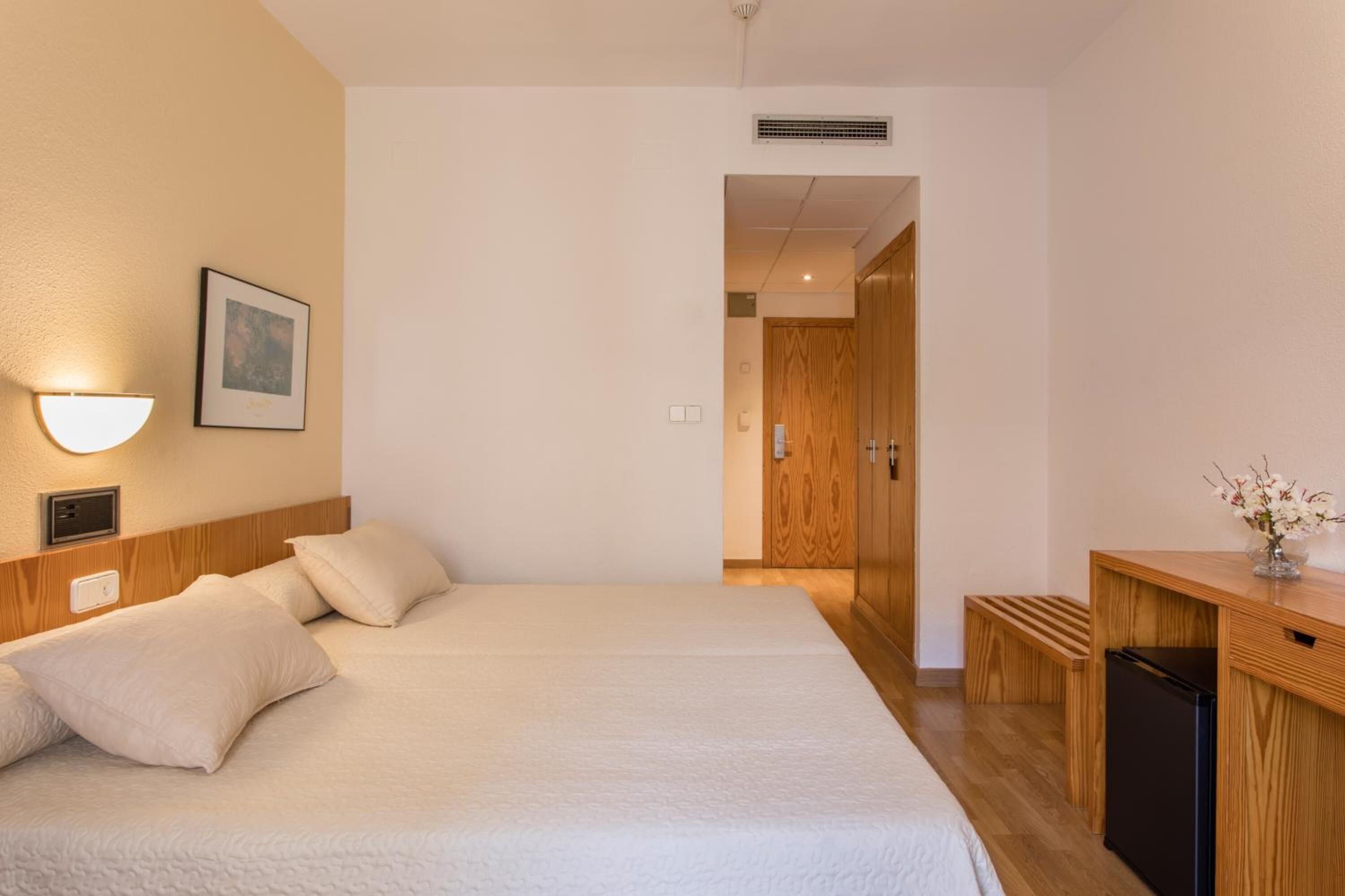 Ein Hotelzimmer mit einem großen Bett, einem Schreibtisch mit Computer, einer Wandlampe und einem gerahmten Bild.