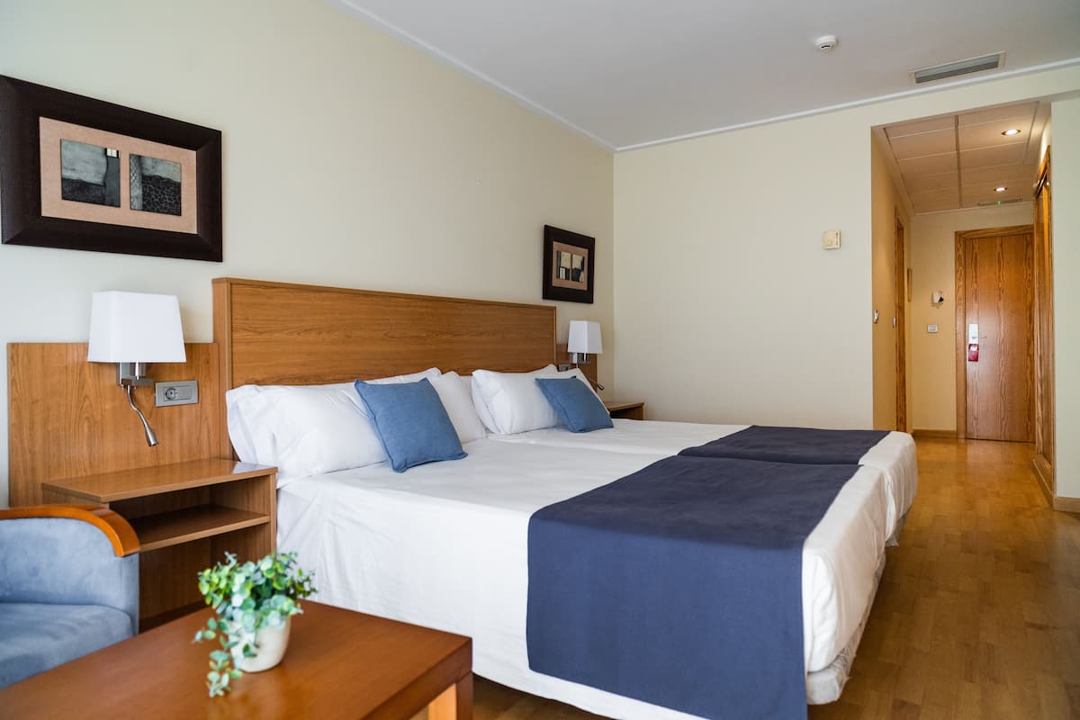 Une chambre d’hôtel avec un grand lit, des tables de chevet, un bureau et une chaise. Il y a deux lampes fixées au mur et une plante en pot sur le bureau.