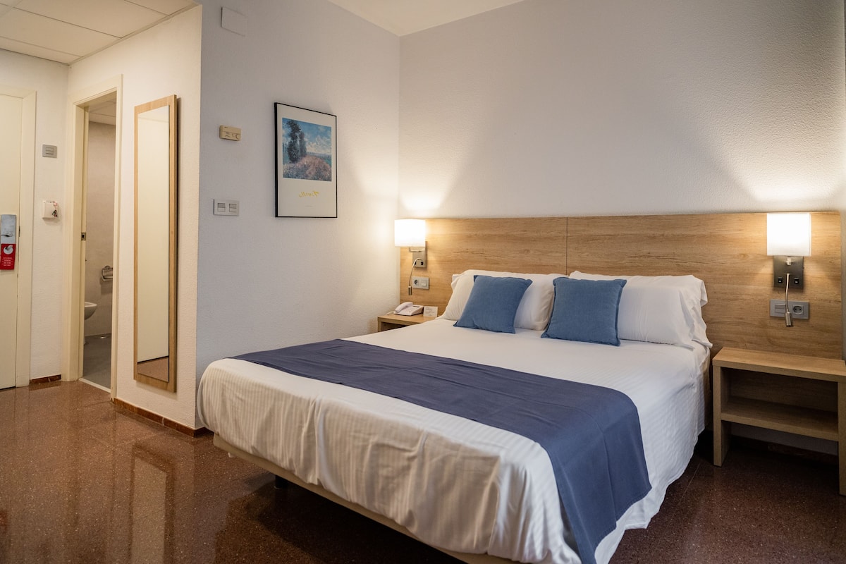 Une chambre d’hôtel comprenant un lit, des tables de chevet, une commode, un miroir, un tableau encadré accroché au mur et une porte donnant sur une autre pièce.