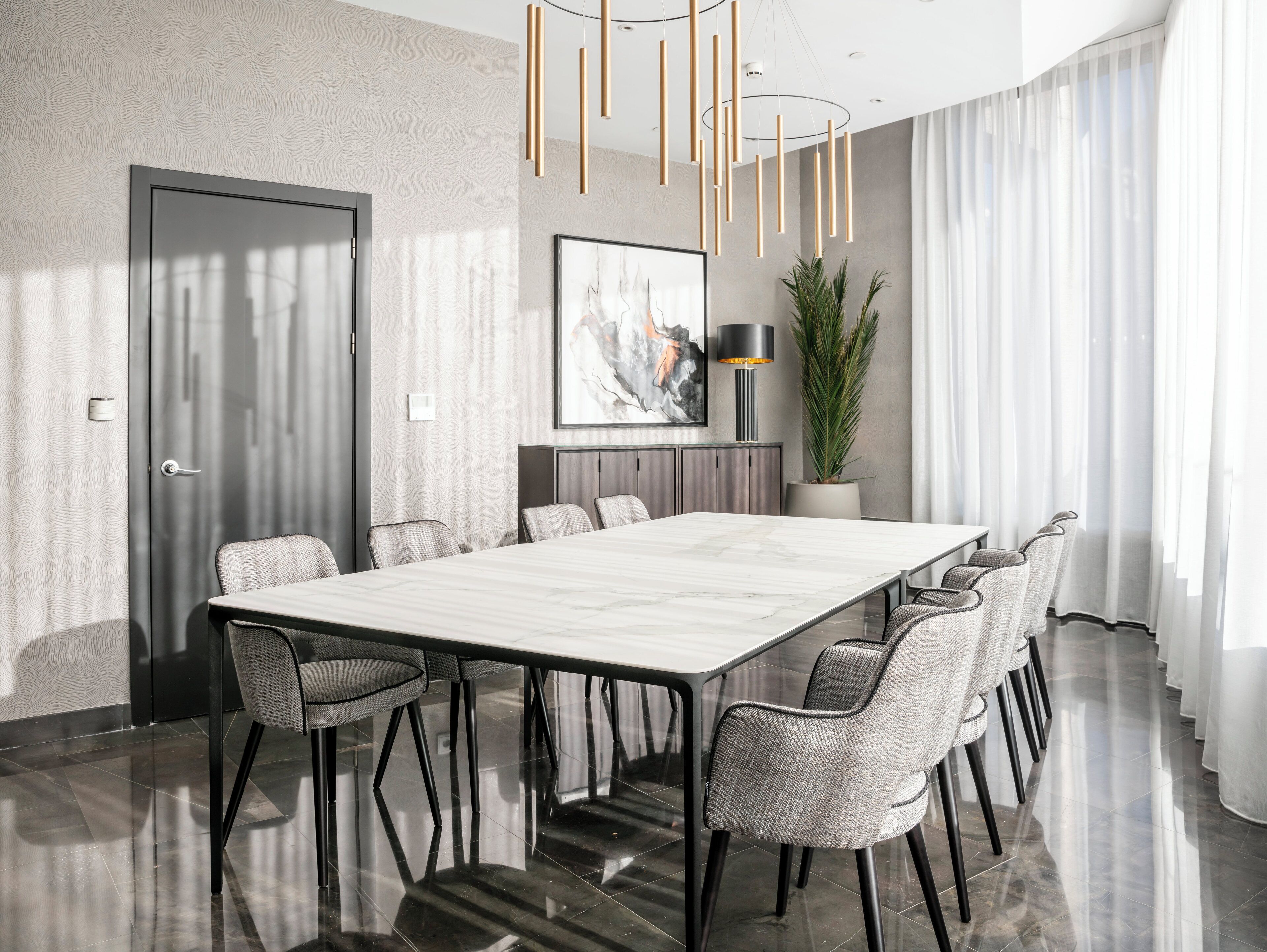 A modern dining room with a marble table, grey upholstered chairs, a potted plant, and a contemporary chandelier.