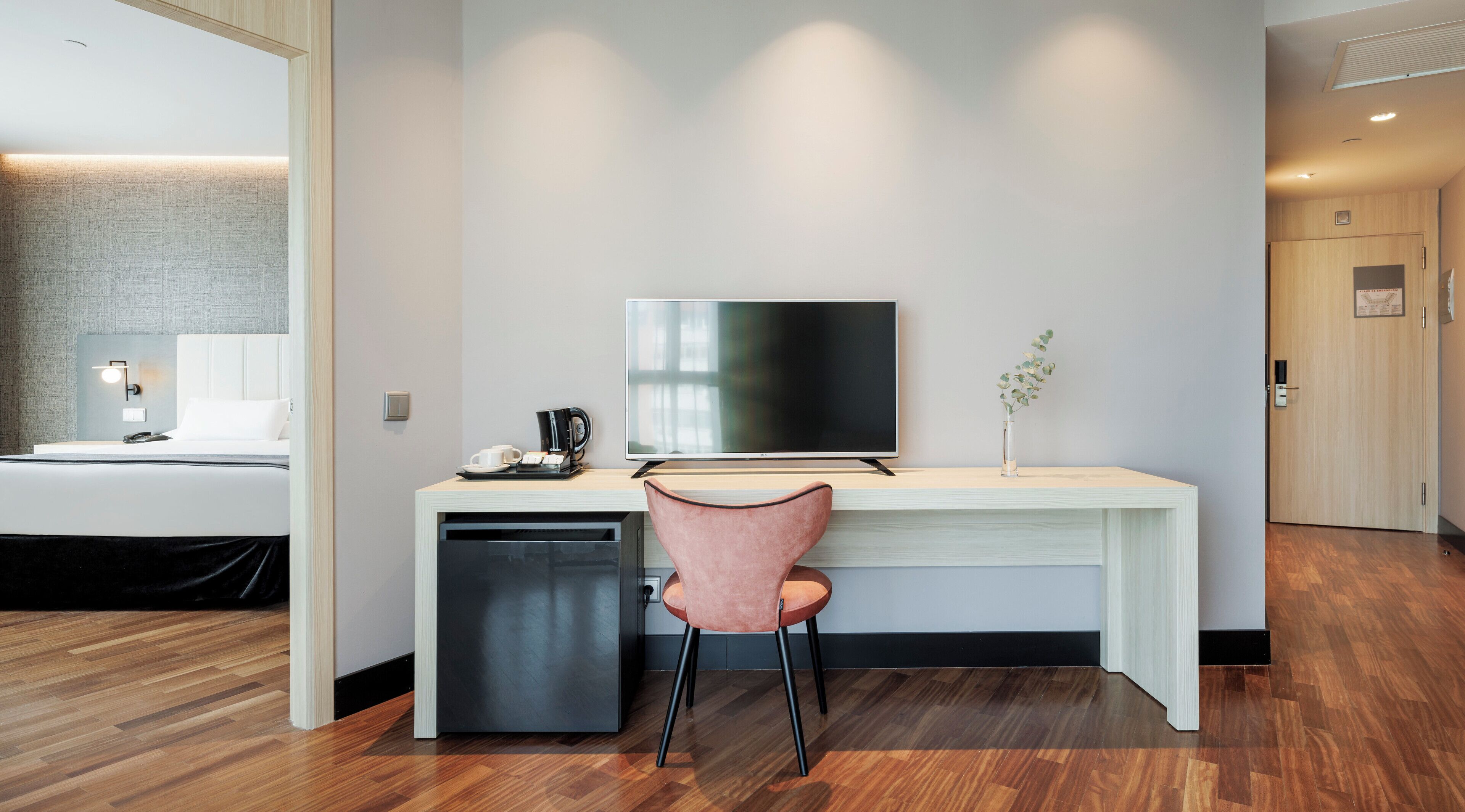 Una habitación de hotel moderna con suelo de madera, un escritorio blanco, un televisor de pantalla plana, una silla rosa, una cafetera y una pequeña planta.