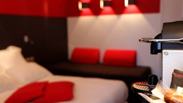 A hotel room with a bed, a desk with a keyboard, and a small table with a cup and a snack.