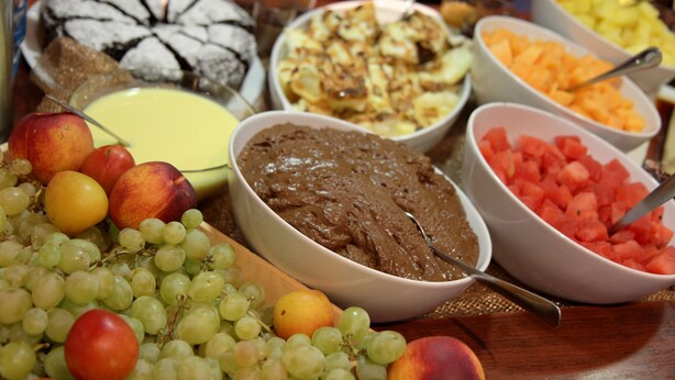 Uma mesa com diversas frutas e sobremesas, incluindo um bolo e tigelas com diferentes tipos de comida.