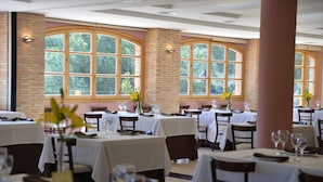 Een restaurant met meerdere tafels, elk voorzien van witte tafelkleden en genummerd servies. Grote ramen met gebogen bovenkanten laten natuurlijk licht de ruimte binnen.