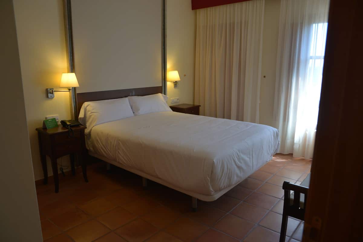 Een hotelkamer met een bed, twee bedlampen, een nachtkastje met een telefoon en een raam met gordijnen.