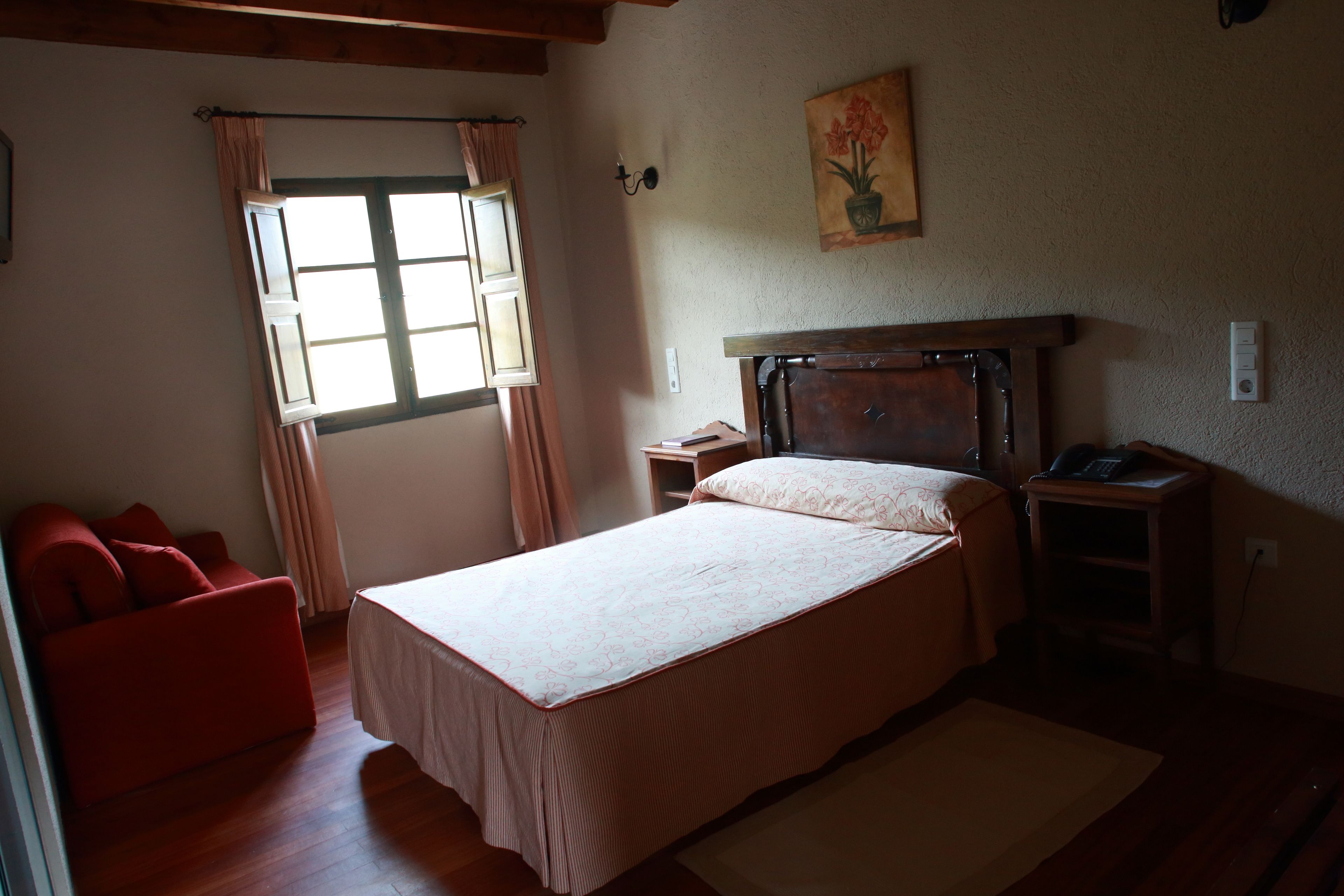 Un dormitorio con cama, mesita de noche, una silla roja, ventana con cortinas y un cuadro en la pared.