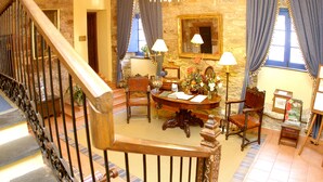 Une vaste pièce dotée d’un escalier en bois, d’une table ronde en bois entourée de chaises, d’un lustre et d’un mur en pierre.