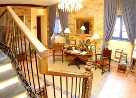 Una sala amplia con una escalera de madera, una mesa redonda de madera y sillas, una araña y una pared de piedra.