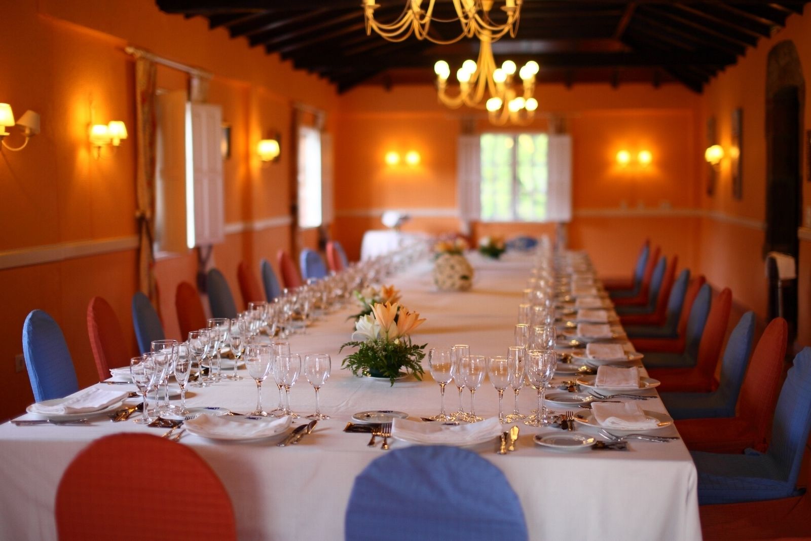 A long dining table set with chairs, glasses, and cutlery, in a room with orange walls and chandeliers.