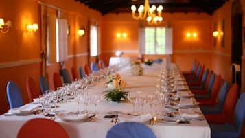 A long dining table set with chairs, glasses, and cutlery, in a room with orange walls and chandeliers.