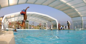 Una piscina interior con techo de cristal, agua transparente y gente nadando y saltando.