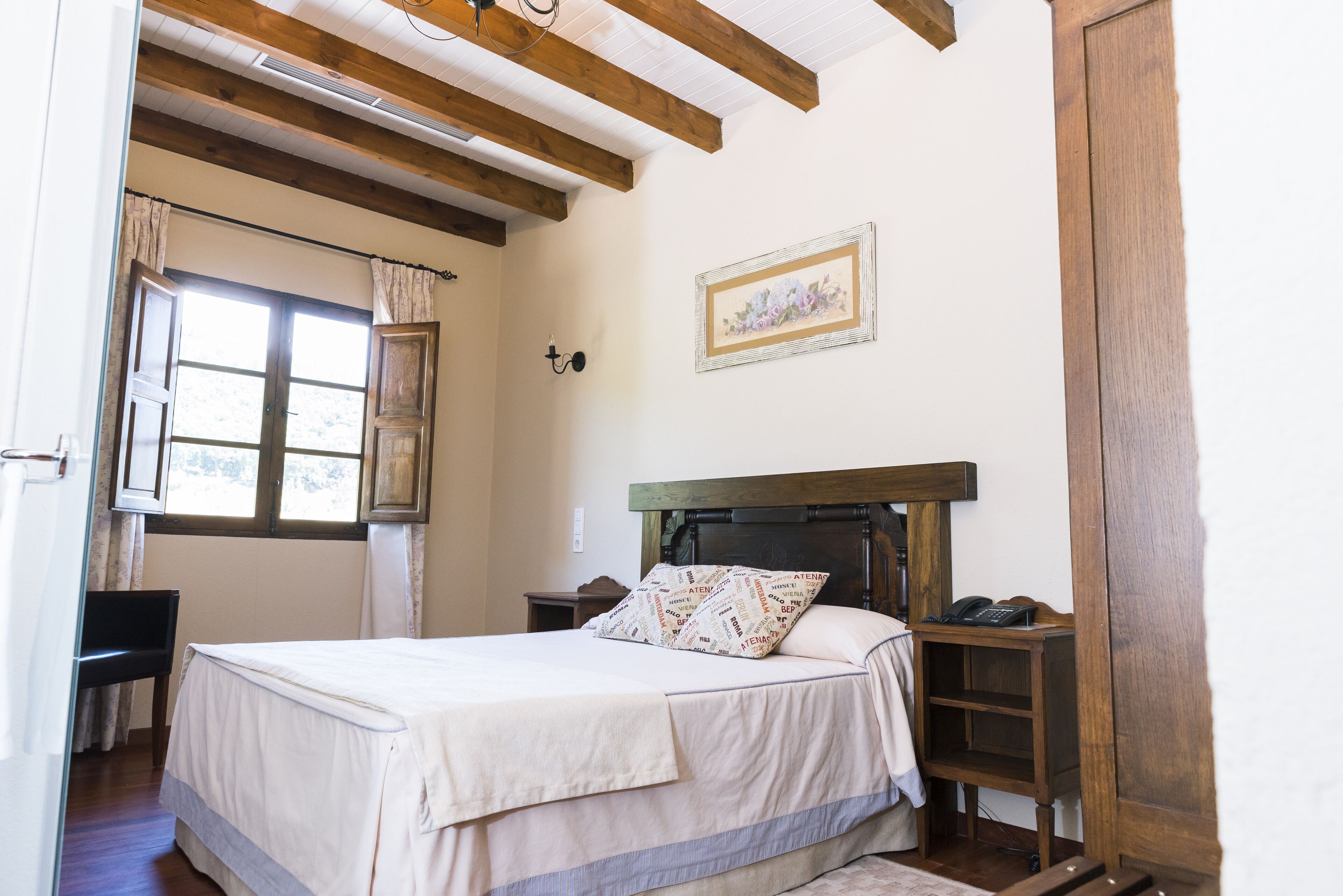 A bedroom with a wooden headboard, a bed with white bedding, a bedside table with a lamp, and a window with curtains.