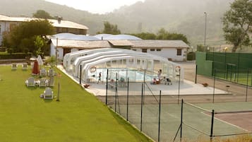A tennis court with a net, a pool, and a covered seating area.
