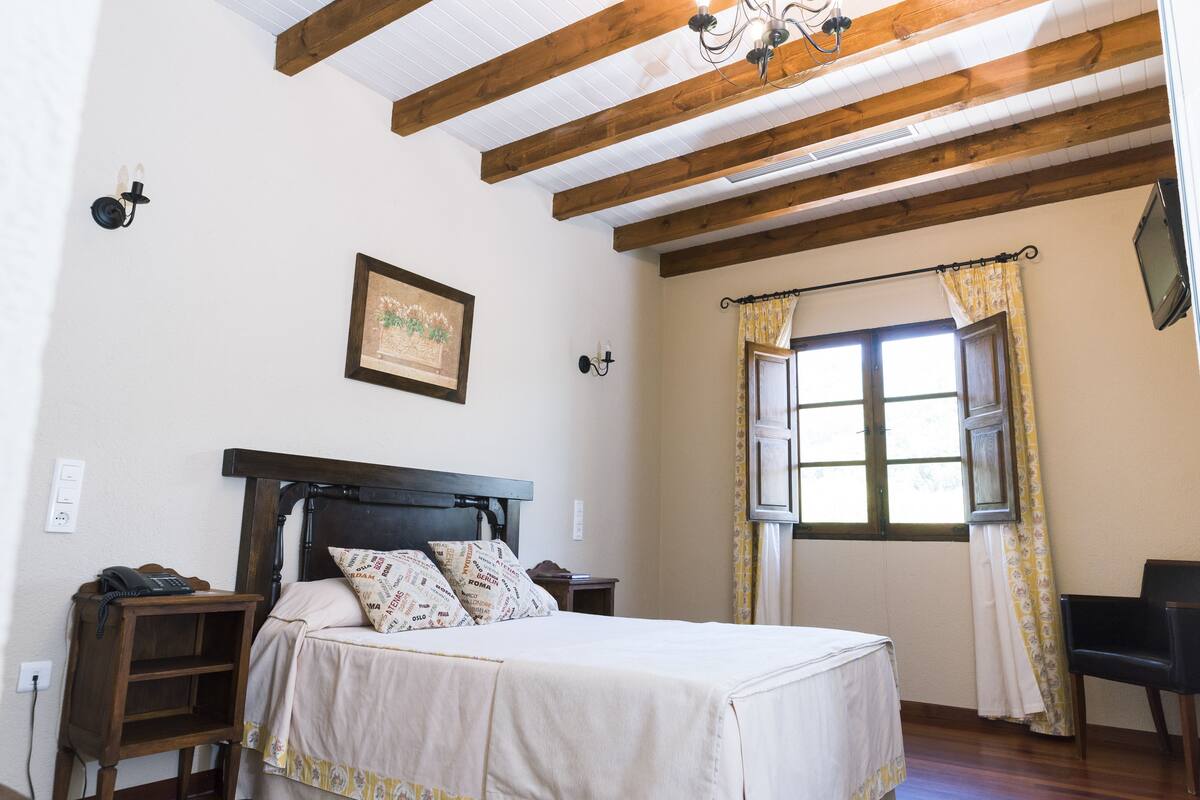 A bedroom with a bed, bedside tables, a nightstand, a framed picture on the wall, a window with curtains, and a chair.