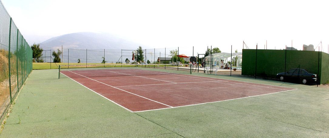 Una cancha de tenis con una cerca verde, superficie de juego roja y una zona circundante verde.
