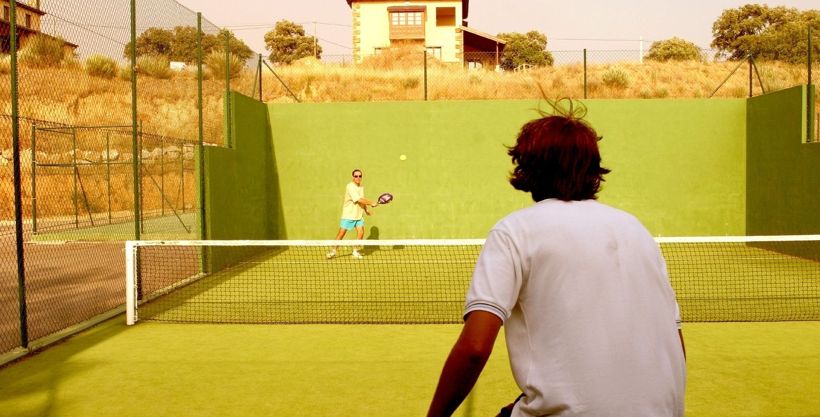 Una cancha de tenis con una red, un jugador con camisa blanca y otro con camisa amarilla.