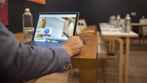 A person using a laptop at a wooden table in a room with a dark wall and several water bottles on a table. - Hotel Fruela (Oviedo)