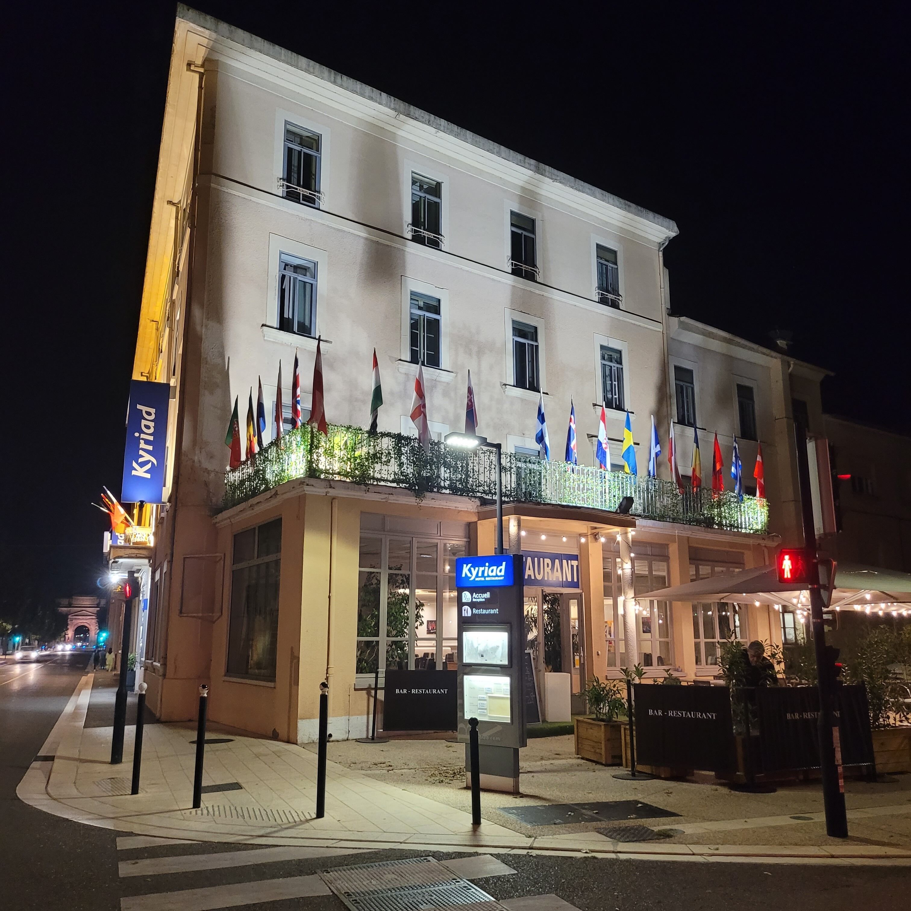 A hotel with multiple flags, a sign for 'Kyriad', and a 'BAR-RESTAURANT' sign.