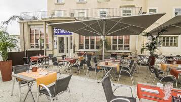 An outdoor cafe with tables and chairs, a sign for a restaurant, and a building in the background.