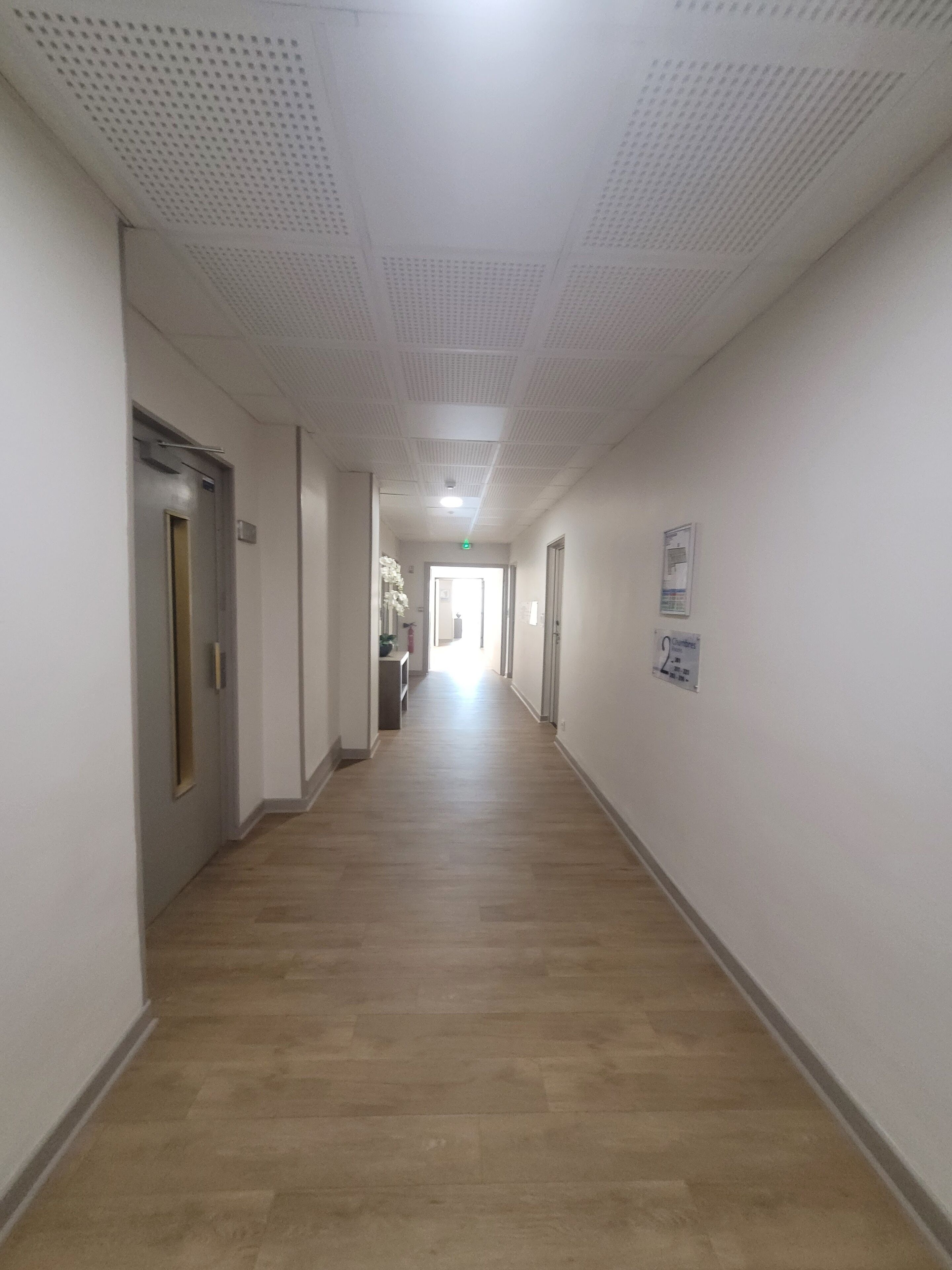 A long, narrow hallway with a light-colored wooden floor and white walls. There are doors on both sides, and a sign on the right wall indicating room numbers.