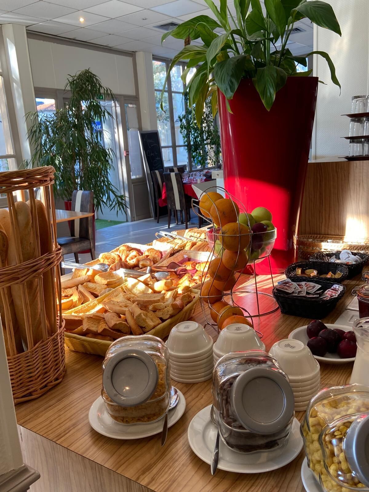 A buffet table with various food items, including bread, fruits, and pastries, in a well-lit indoor setting with large windows and potted plants.