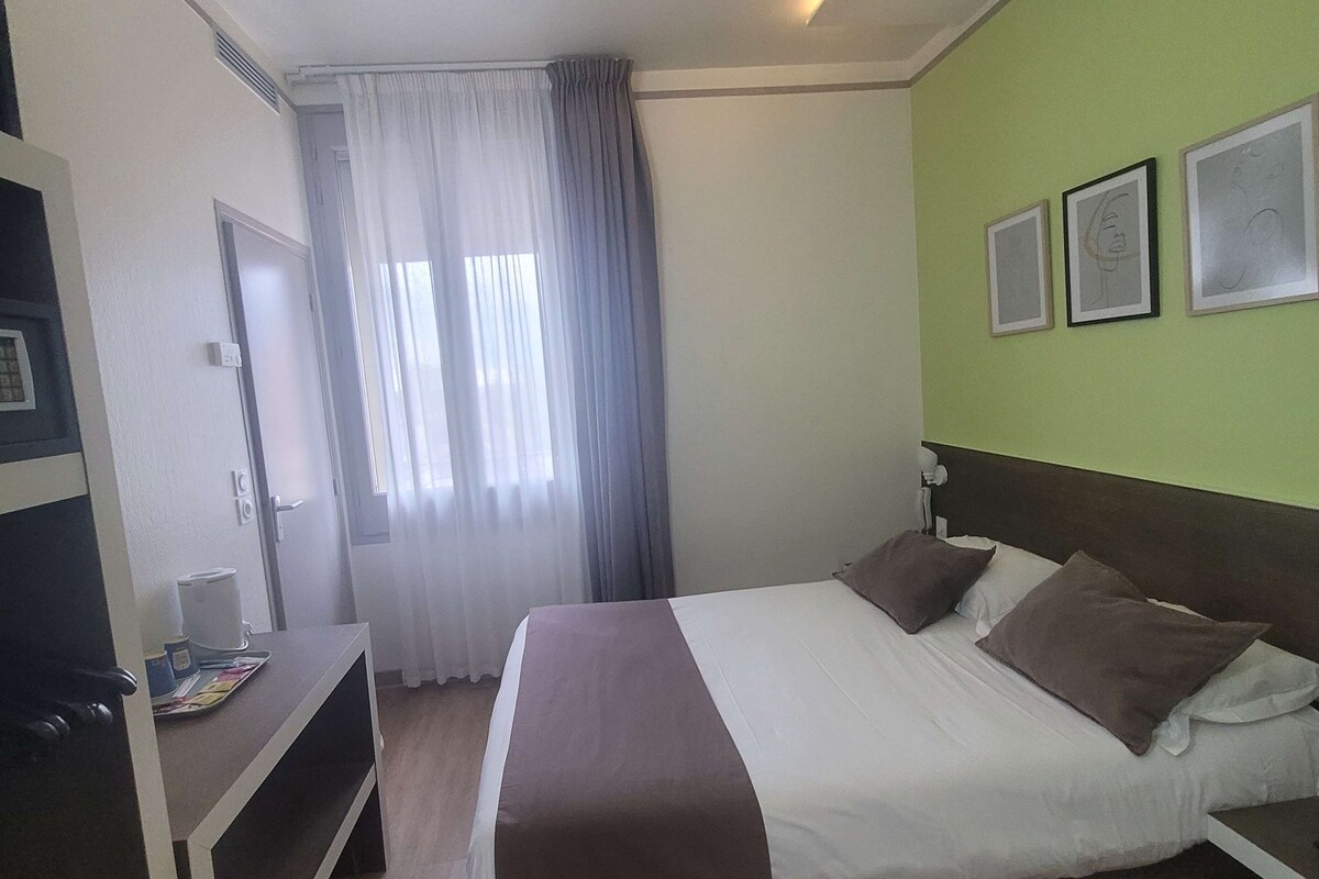 Habitación de hotel con cama, escritorio, silla, ventana con cortinas y tres cuadros en la pared.