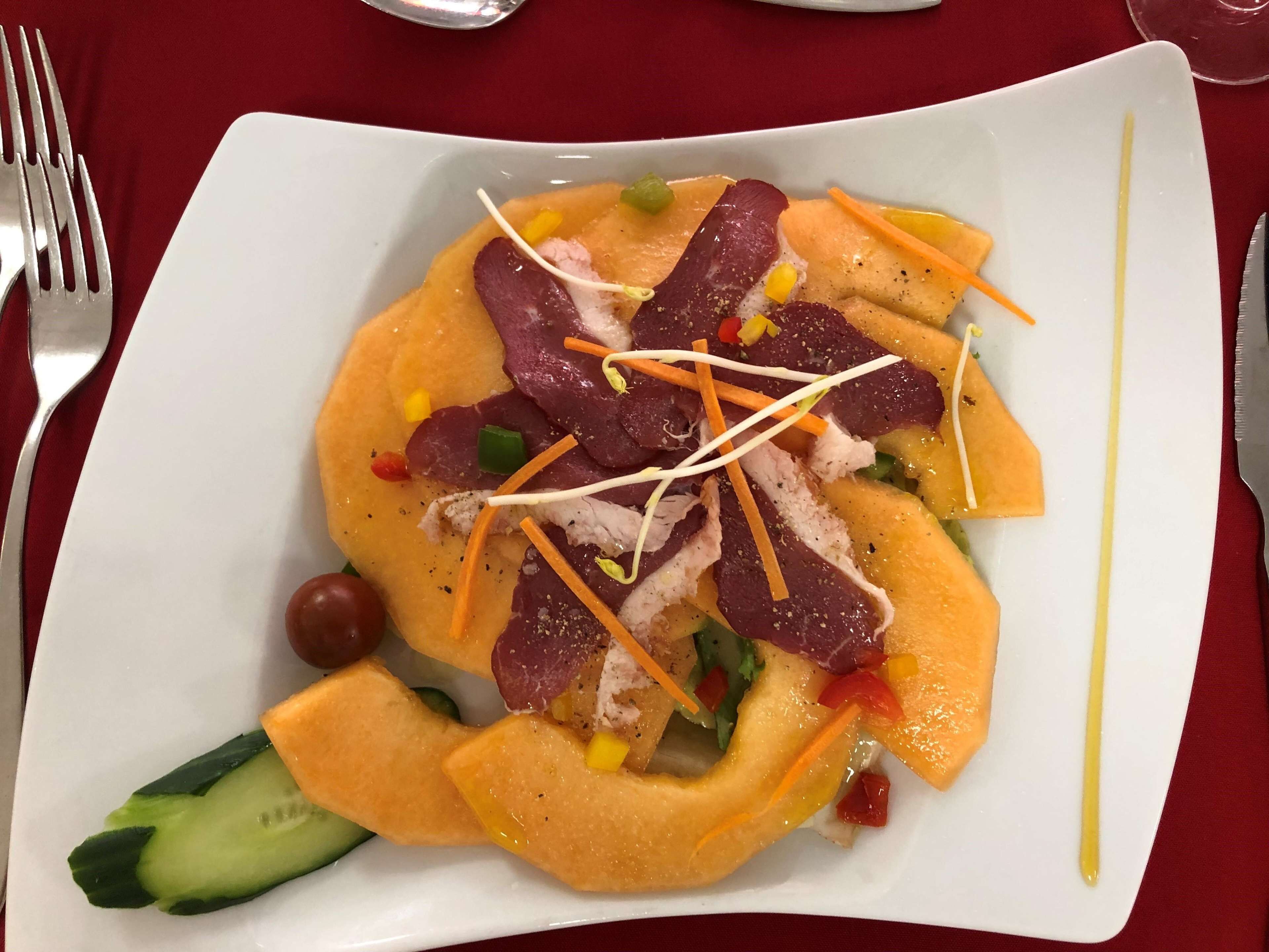 Ein weißer Teller mit einem Salat aus Melonenstücken, Karotten und Fleisch, auf einer roten Tischdecke.