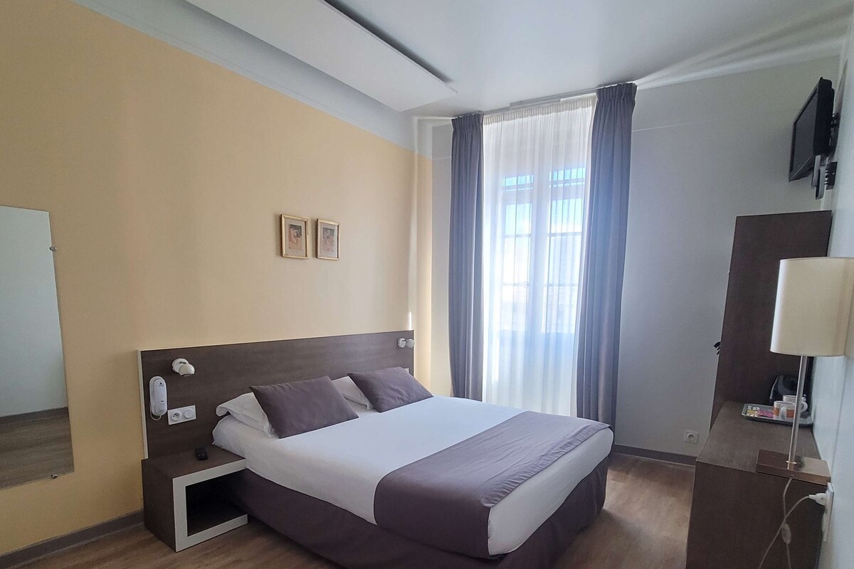 Una habitación de hotel con una cama grande, una mesita de noche con teléfono, un escritorio con lámpara y un televisor en la pared.