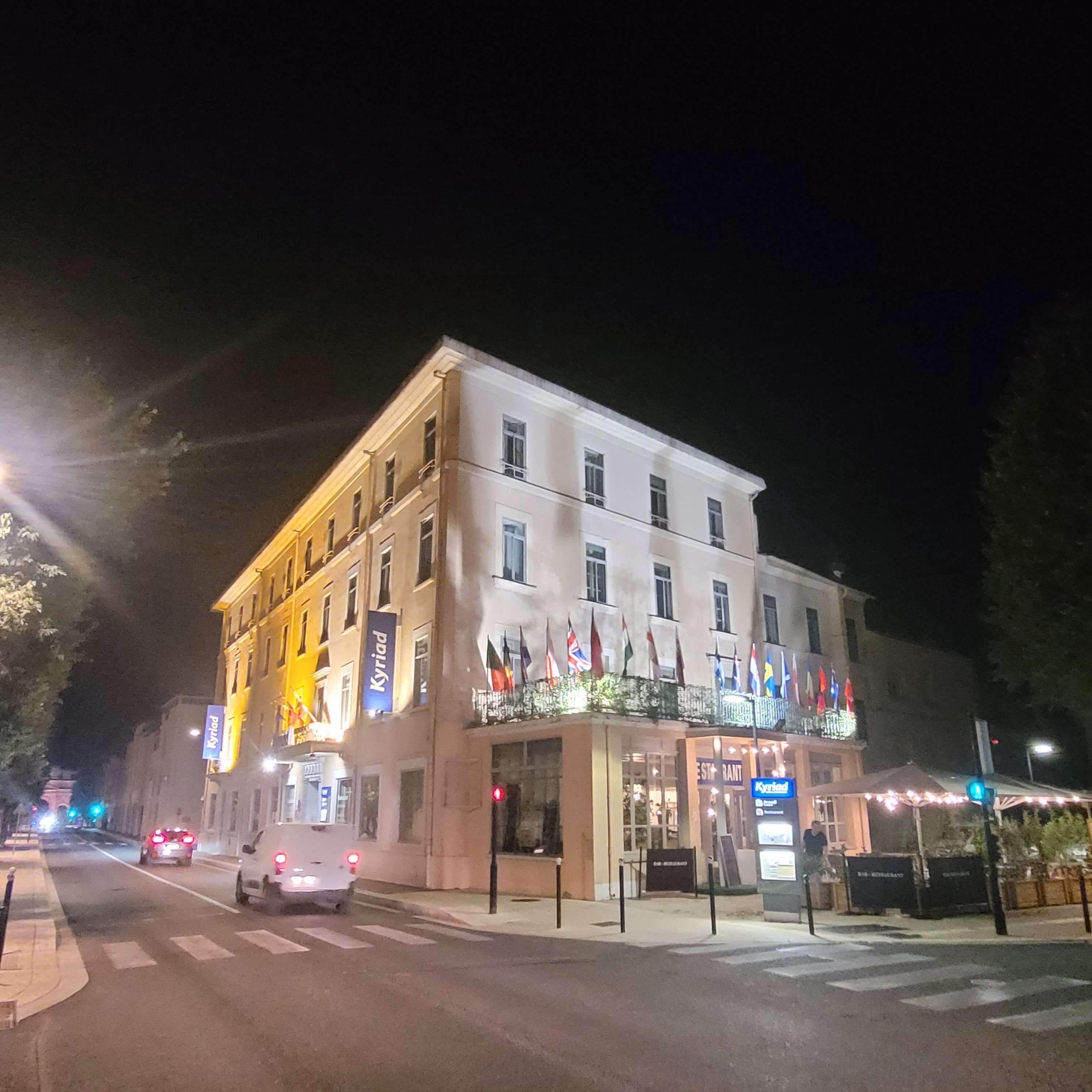 A multi-story hotel with a restaurant on the ground floor, flags on the balcony, and a pedestrian crossing in front.