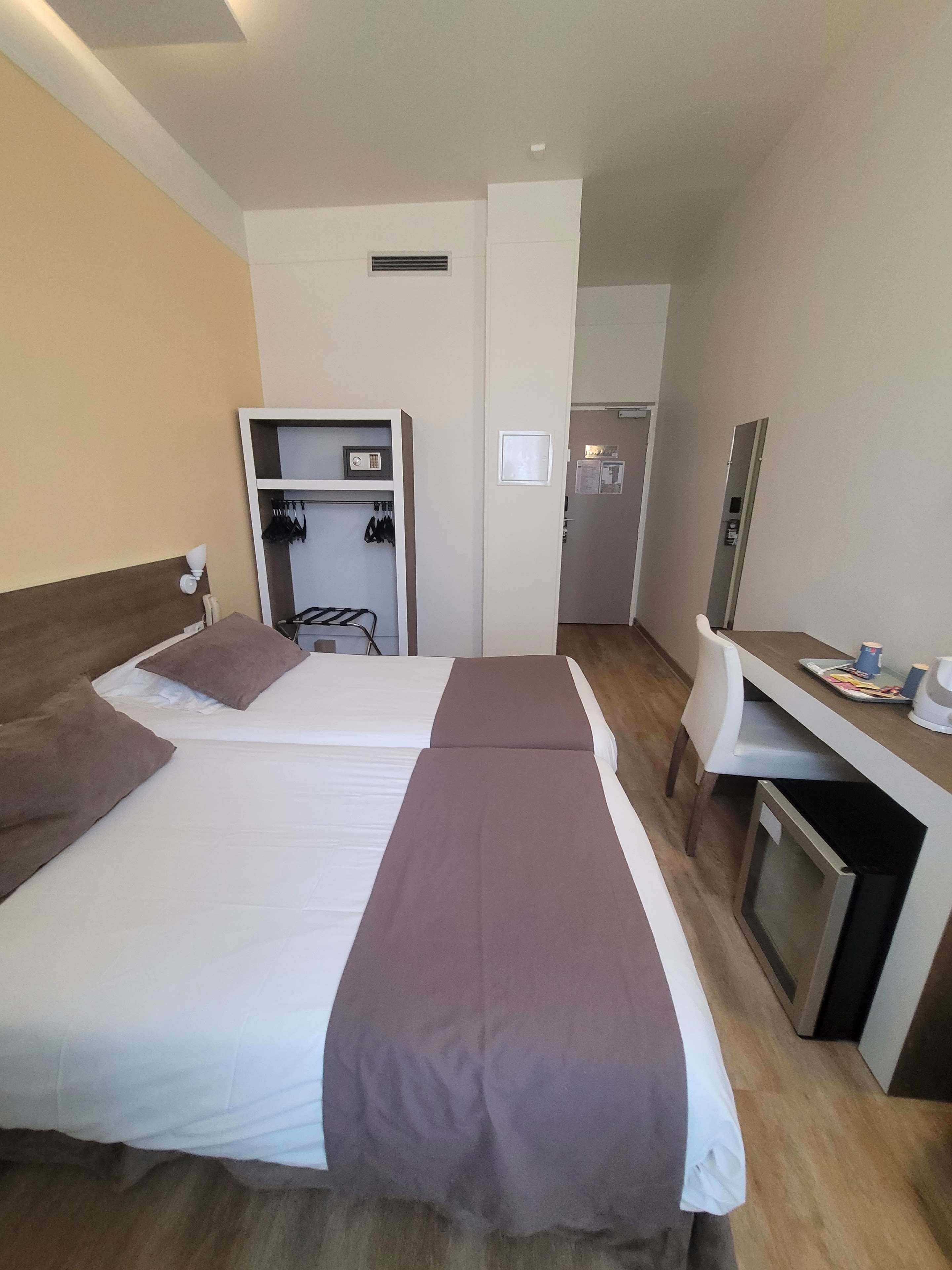 A hotel room with two beds, a desk, a mirror, a wardrobe, and a small kitchenette.