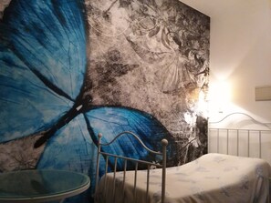 Une chambre avec un grand motif de papillons peint sur le mur, un lit recouvert d’une couvre-lit à motifs floraux et une table de chevet avec une lampe.