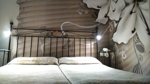 Un lit avec une tête de lit en métal, un mur recouvert d’un papier peint à motifs floraux et une table de chevet avec une lampe.