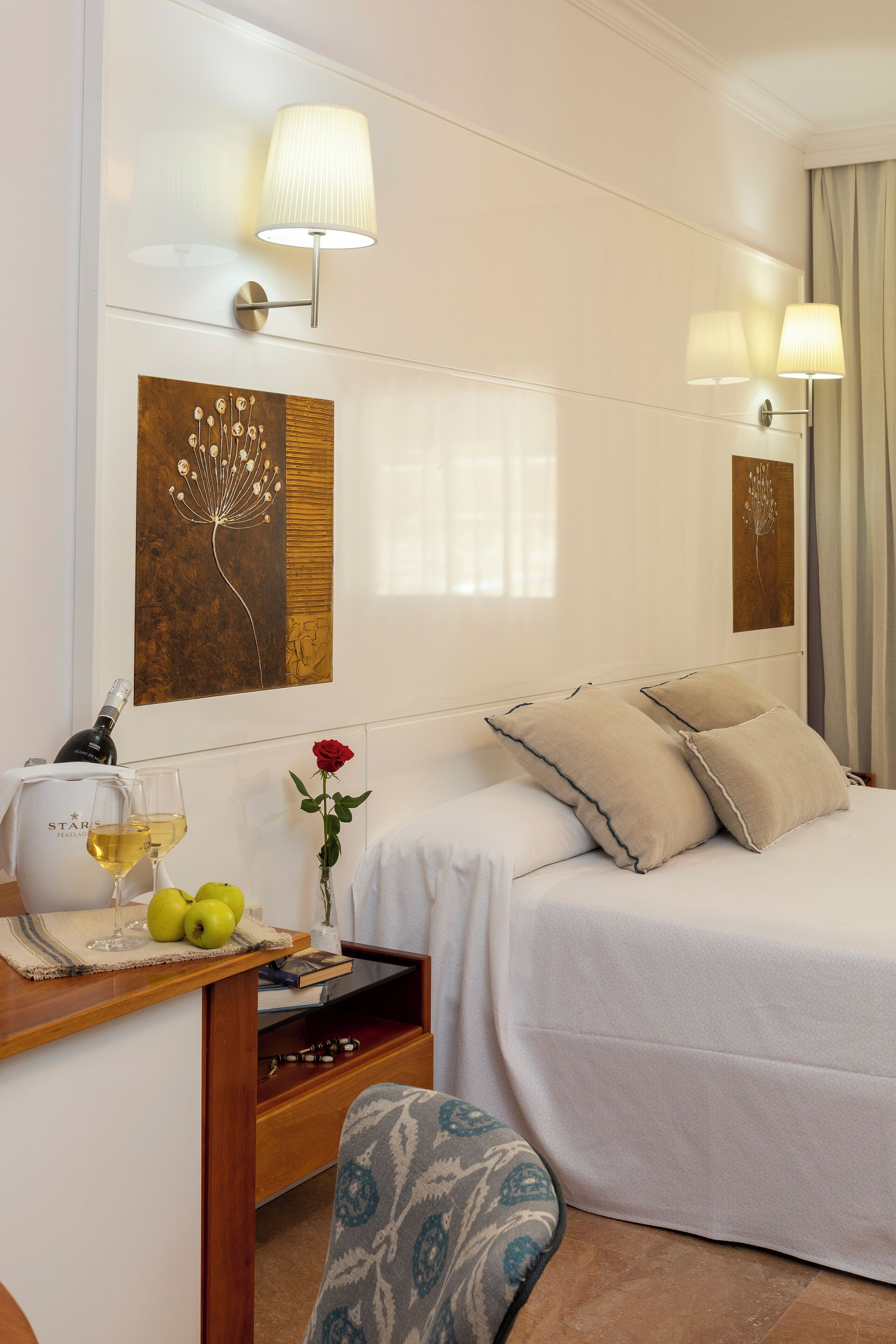A hotel room with a bed, bedside lamps, a table with a bottle of wine, glasses, and apples.