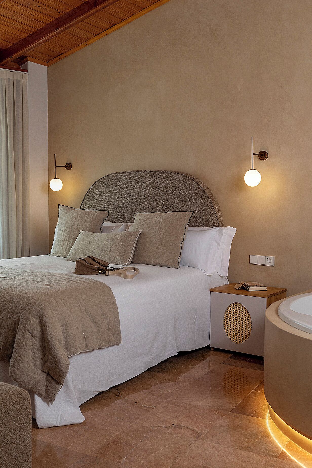 Un dormitorio con cama, mesitas de noche, lámpara de pared y baño con lavabo y espejo.