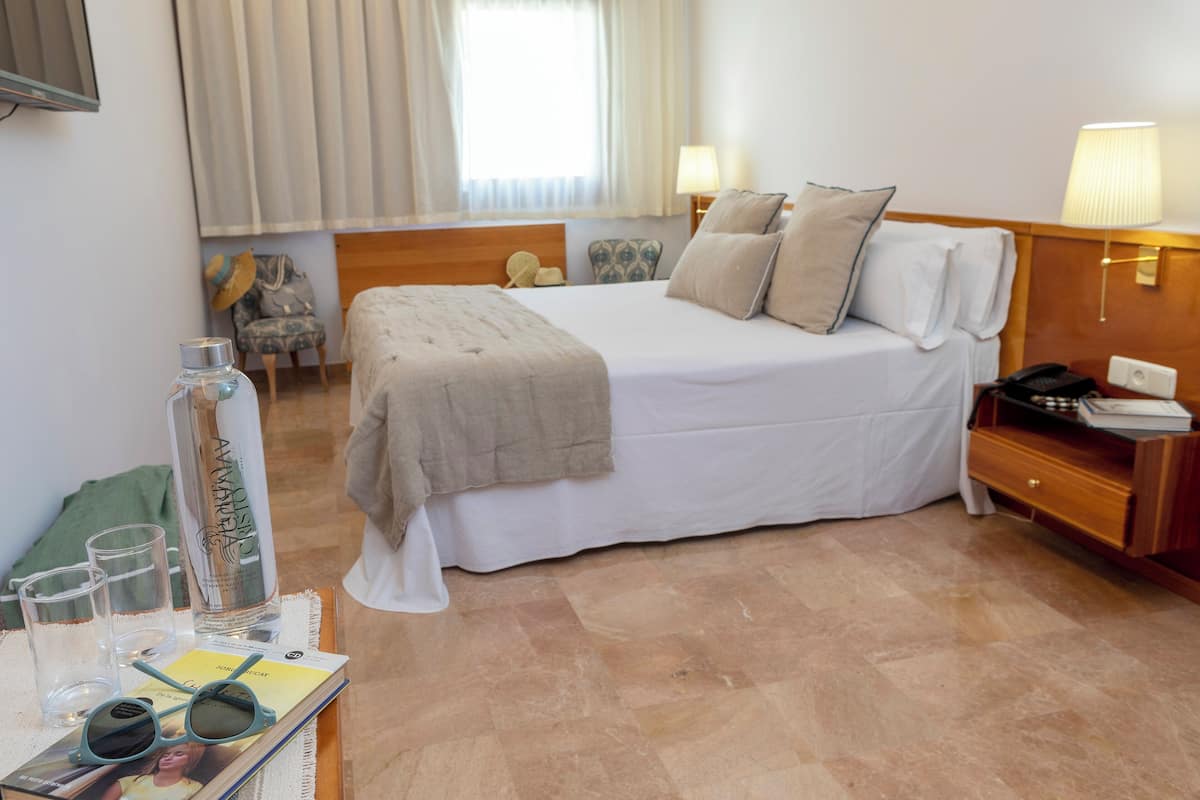 Une chambre d’hôtel avec un lit, des tables de chevet, une lampe, une chaise et une fenêtre avec des rideaux.