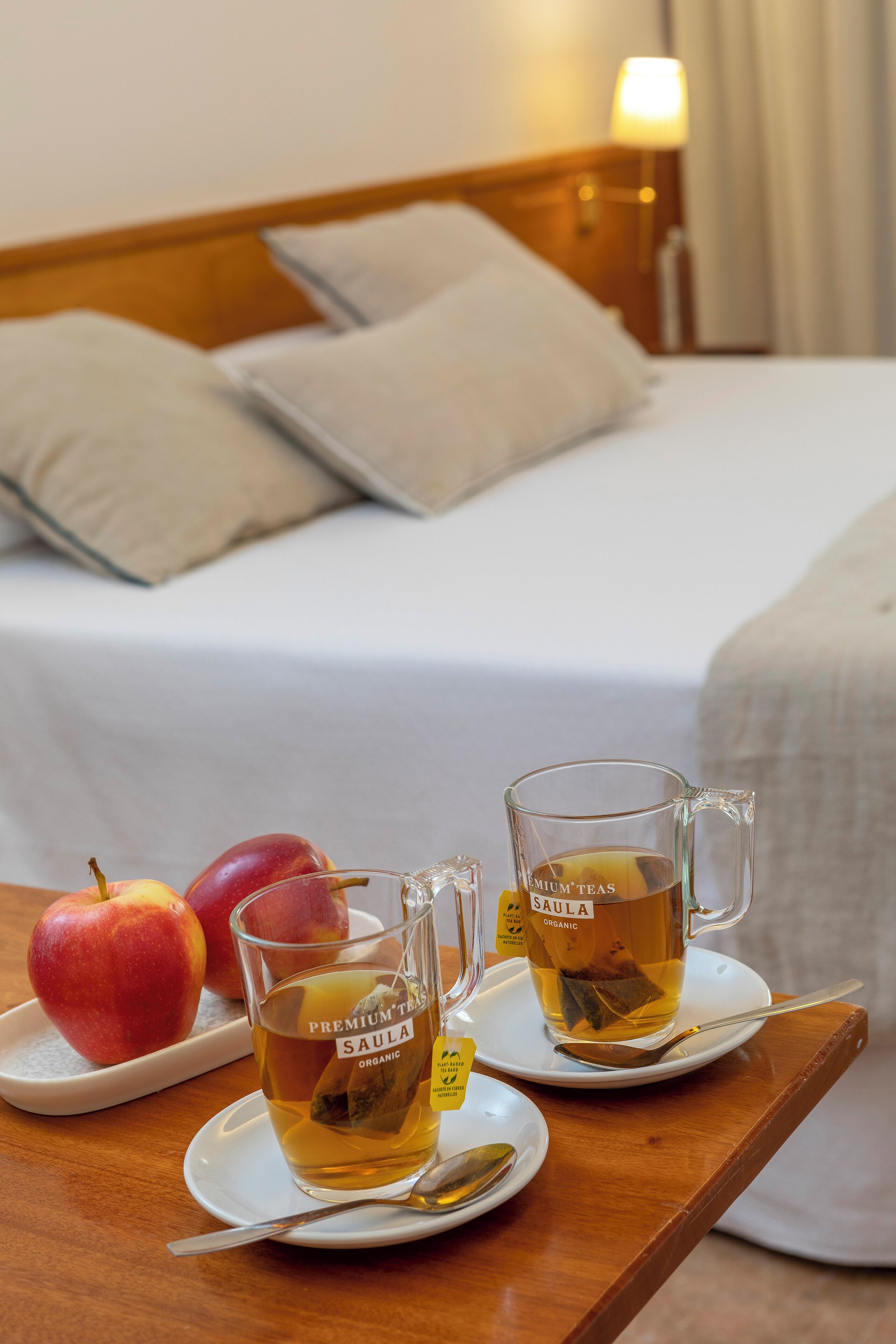 A neatly made bed with pillows, a bedside table with a lamp, and a tray with two glasses of tea and apples.