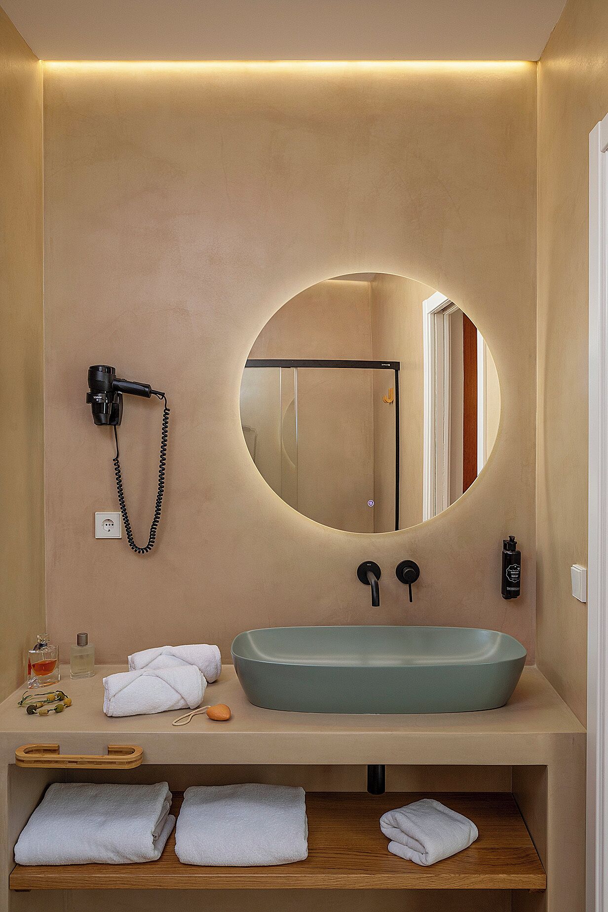 A modern bathroom with a large oval mirror, a beige vanity, a hairdryer, and neatly folded towels.
