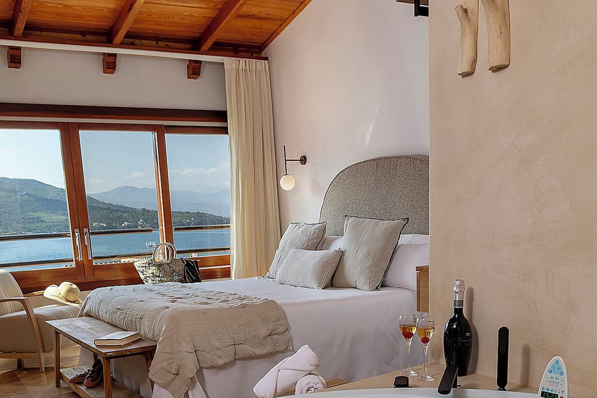 Une chambre avec une grande baignoire, un lit avec des oreillers, une table de chevet avec un livre et une bouteille, et une vue sur la mer par la fenêtre.