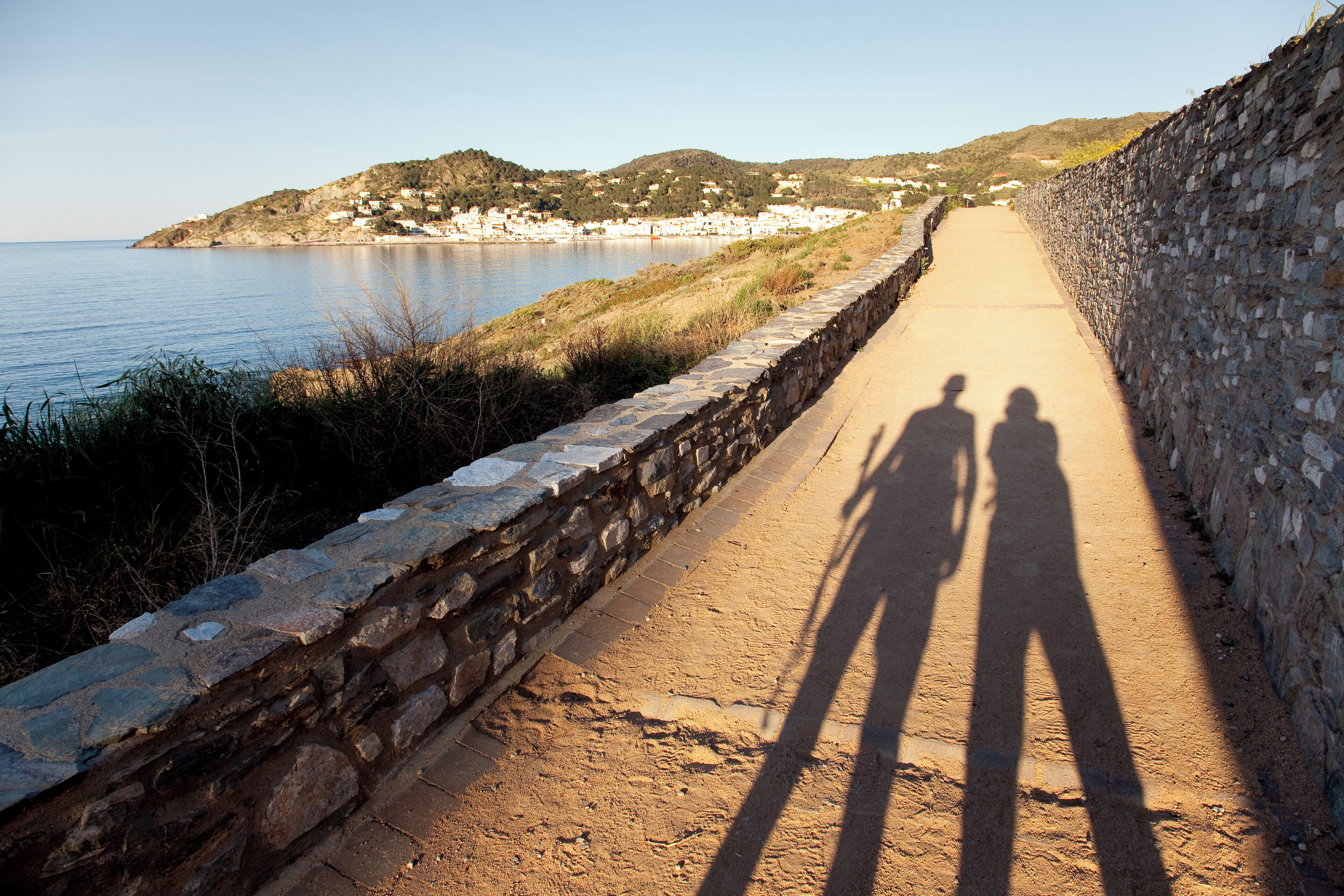 A coastal road with stone walls, a clear sky, and a distant view of houses and hills.