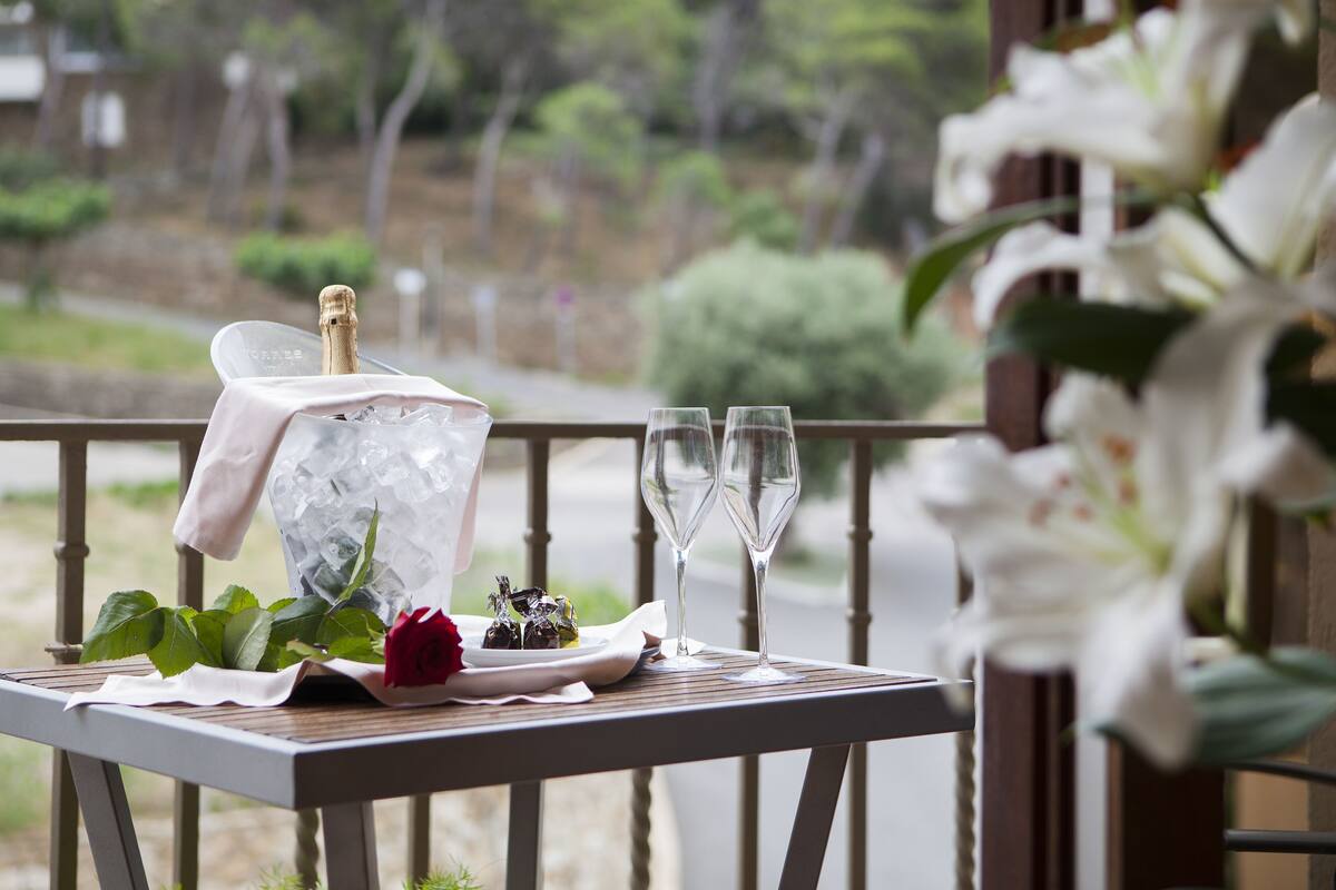Une table avec une bouteille de champagne, deux verres, une rose et une assiette de nourriture, le tout placé sur un balcon doté d’une balustrade et avec de la verdure en arrière-plan.