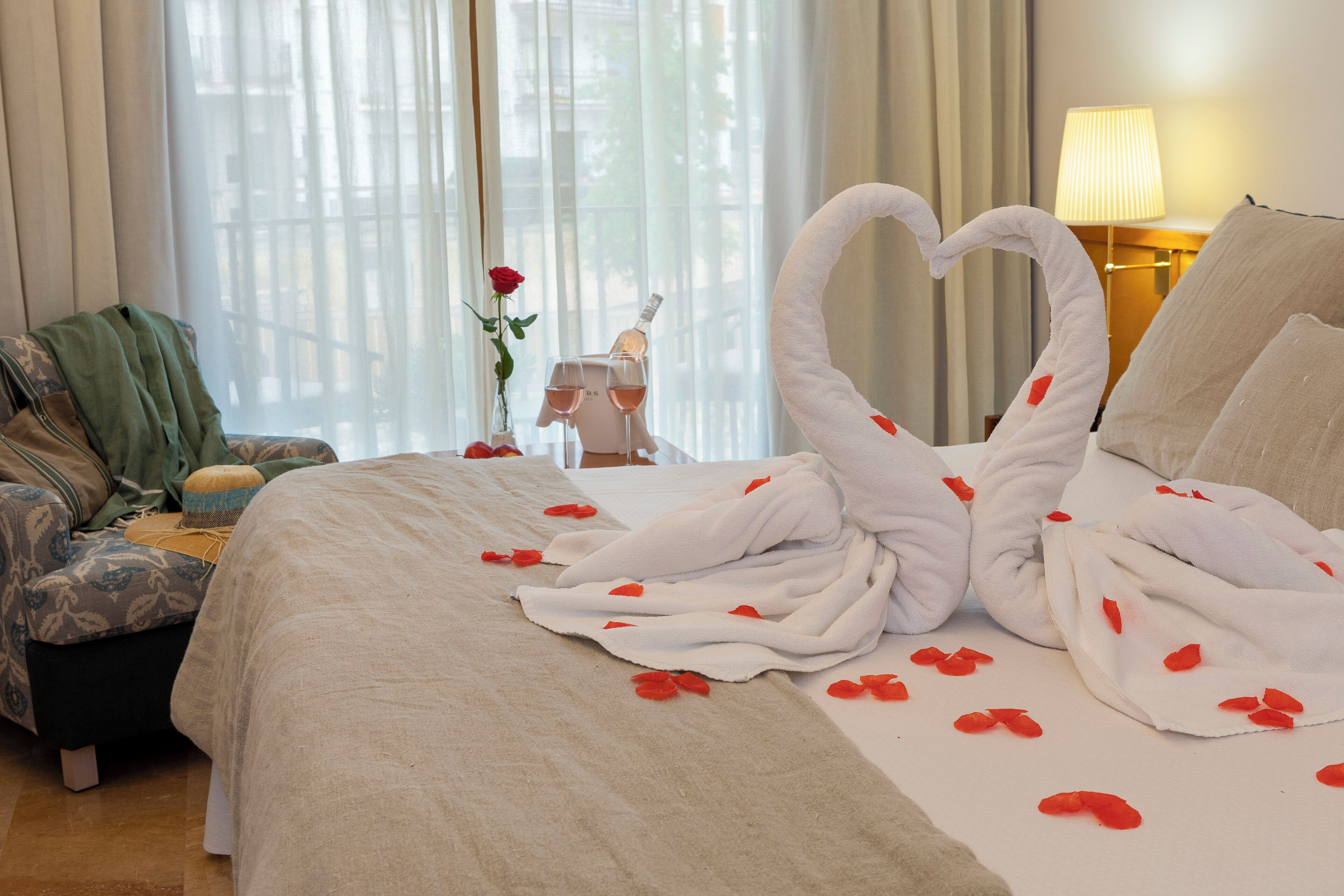 A hotel room with a bed adorned with a swan-shaped arrangement of towels and rose petals, a vase with a rose, and a bottle of champagne.