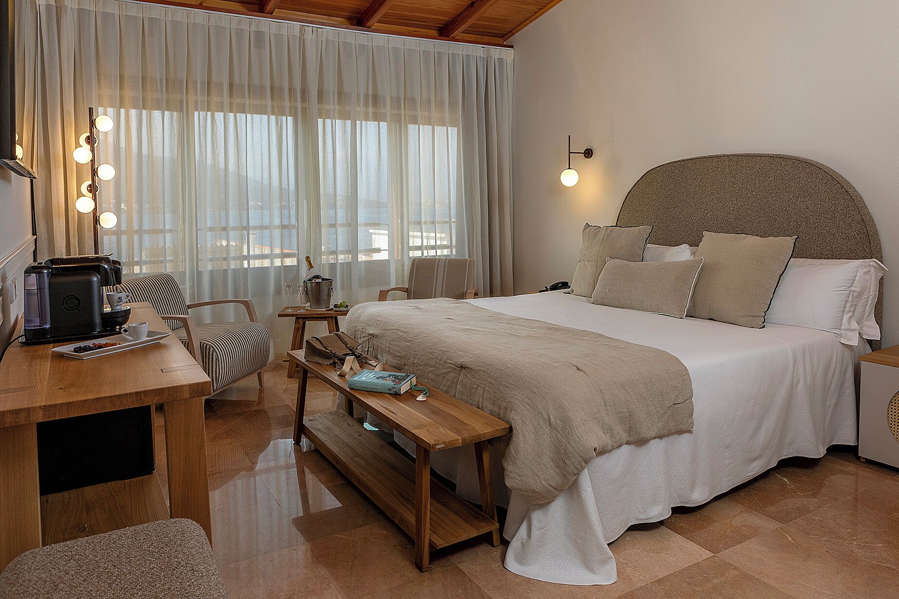 Habitación de hotel con cama, mesita de noche, escritorio con cafetera, una silla y ventana con cortinas.