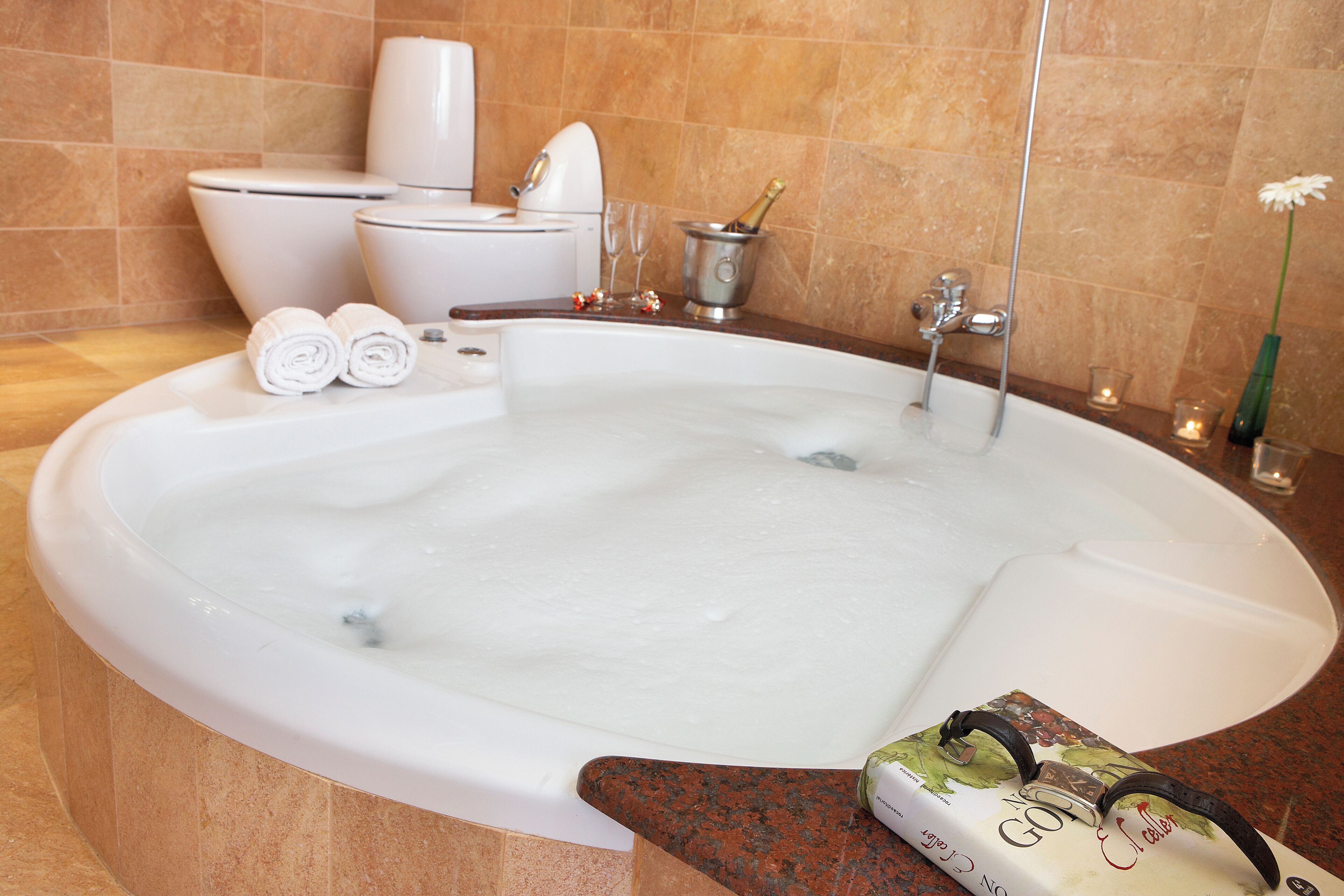 A large bathtub with a bottle of champagne, two rolled towels, and a book on the edge.
