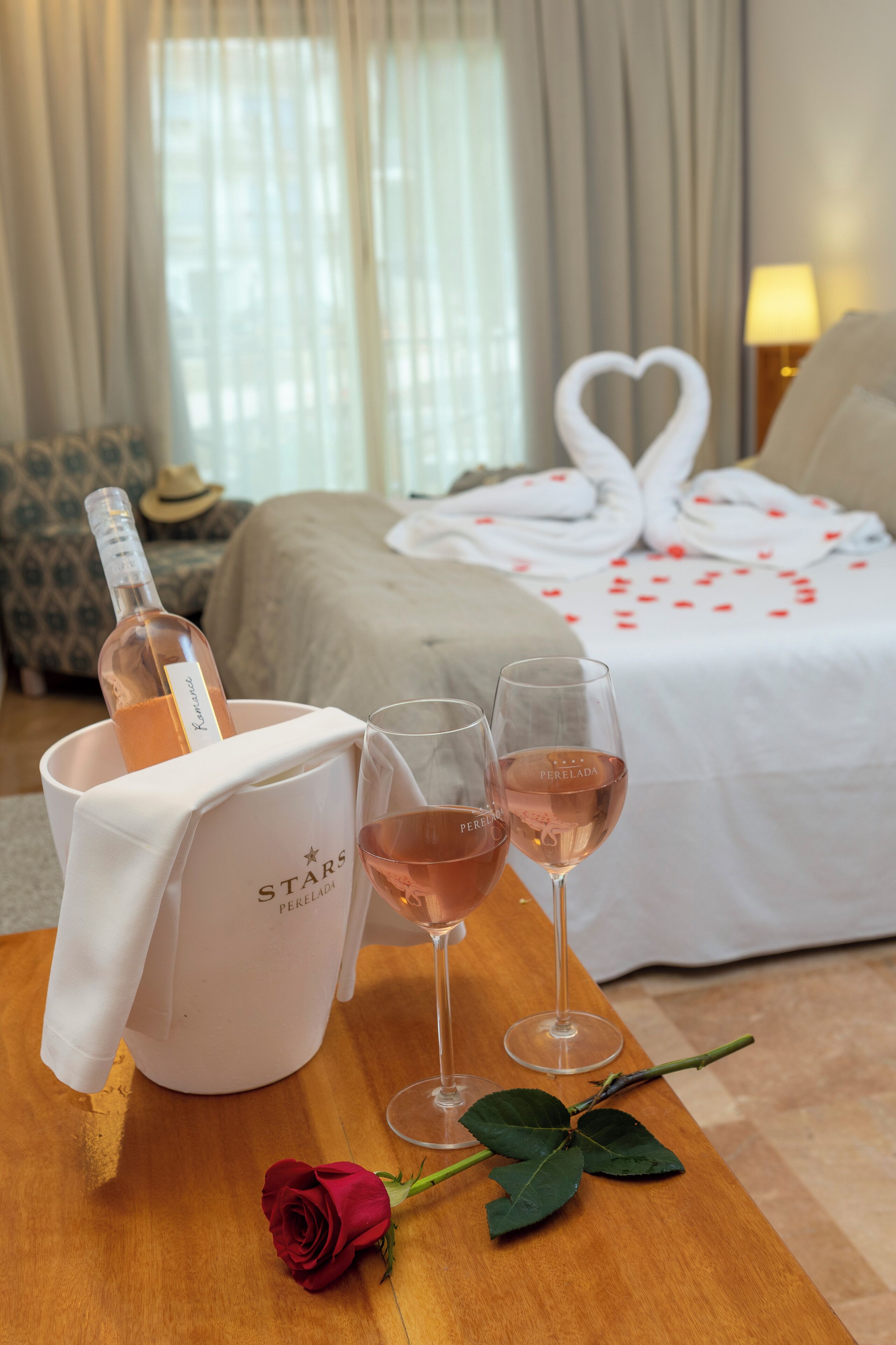 Una habitación de hotel con una cama, una mesita de noche con una botella de vino y copas, una rosa, y una disposición de toallas con forma de corazón.