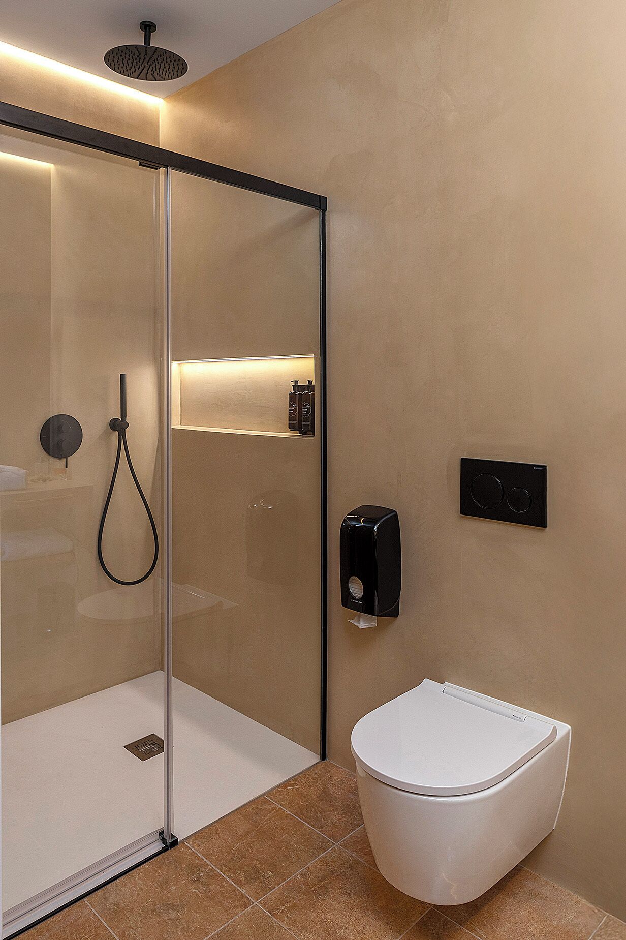 A modern bathroom with a glass-enclosed shower, a toilet, and a soap dispenser on the wall.