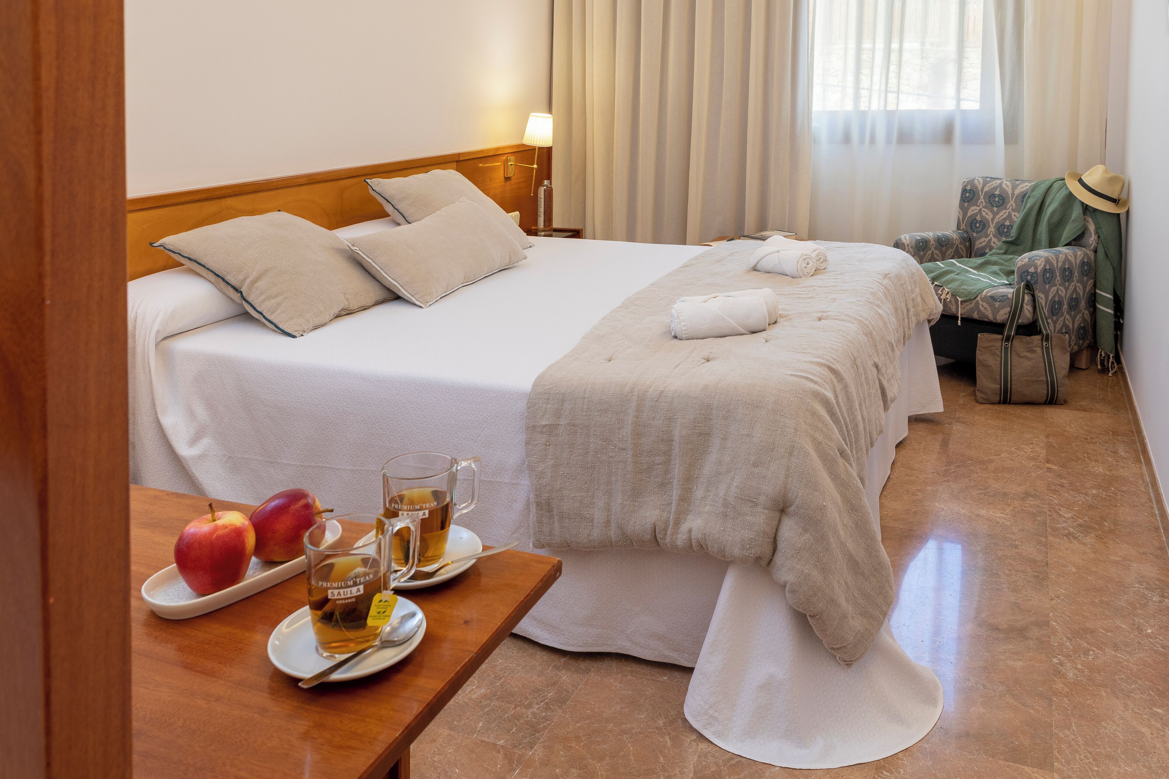 Una habitación de hotel con una cama bien hecha, una mesa con manzanas y té, y una silla con un sombrero.